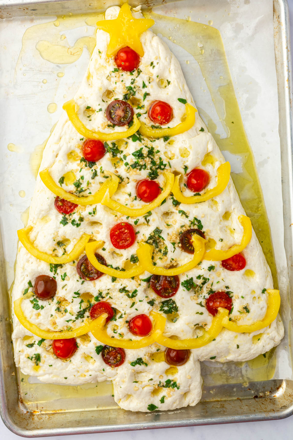 Raw Christmas tree focaccia decorated with yellow pepper star and garland, cherry tomatoes, and fresh parsley ready to bake on sheet pan.