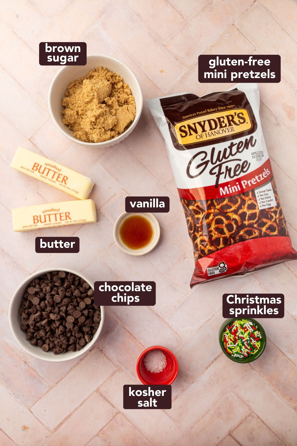 Ingredients for gluten-free Christmas pretzel bark including Snyder's gluten-free mini pretzels, unsalted butter, brown sugar, vanilla extract, semisweet chocolate chips, kosher salt, and holiday sprinkles arranged on white surface.