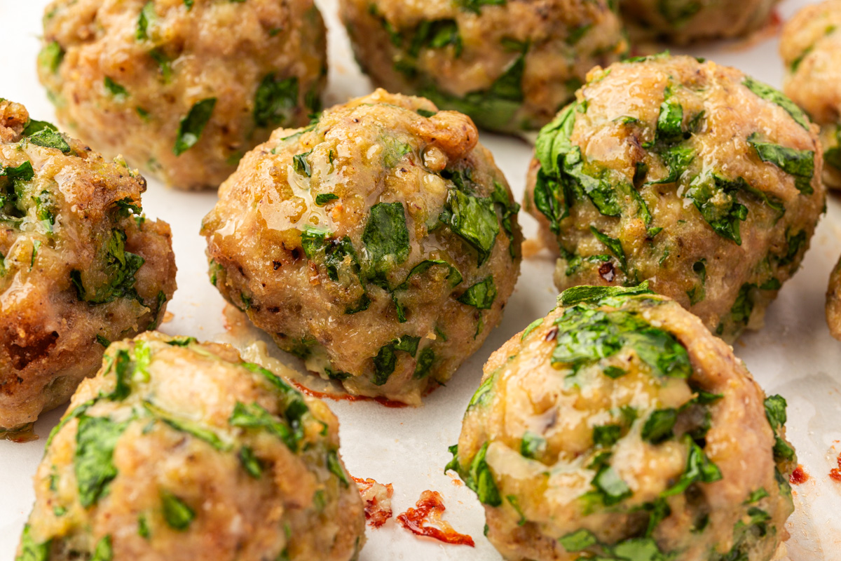 Close-up of several baked meatballs with visible chopped green herbs and small onion pieces, resting on a white surface. The meatballs appear juicy and lightly browned.
