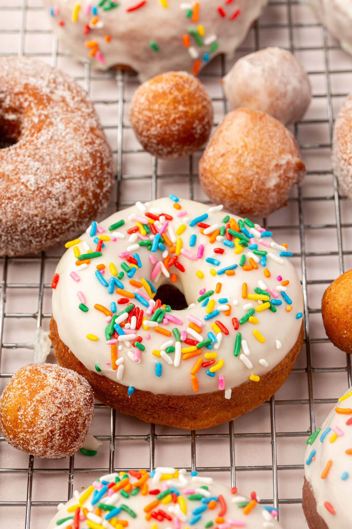 A vanilla-glazed donut with colorful sprinkles sits on a cooling rack surrounded by sugared donuts and donut holes.