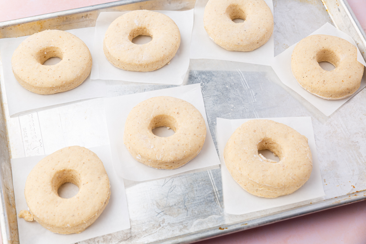 Seven unbaked, round doughnuts with holes in the center rest on squares of parchment paper on a metal baking sheet, ready for baking.