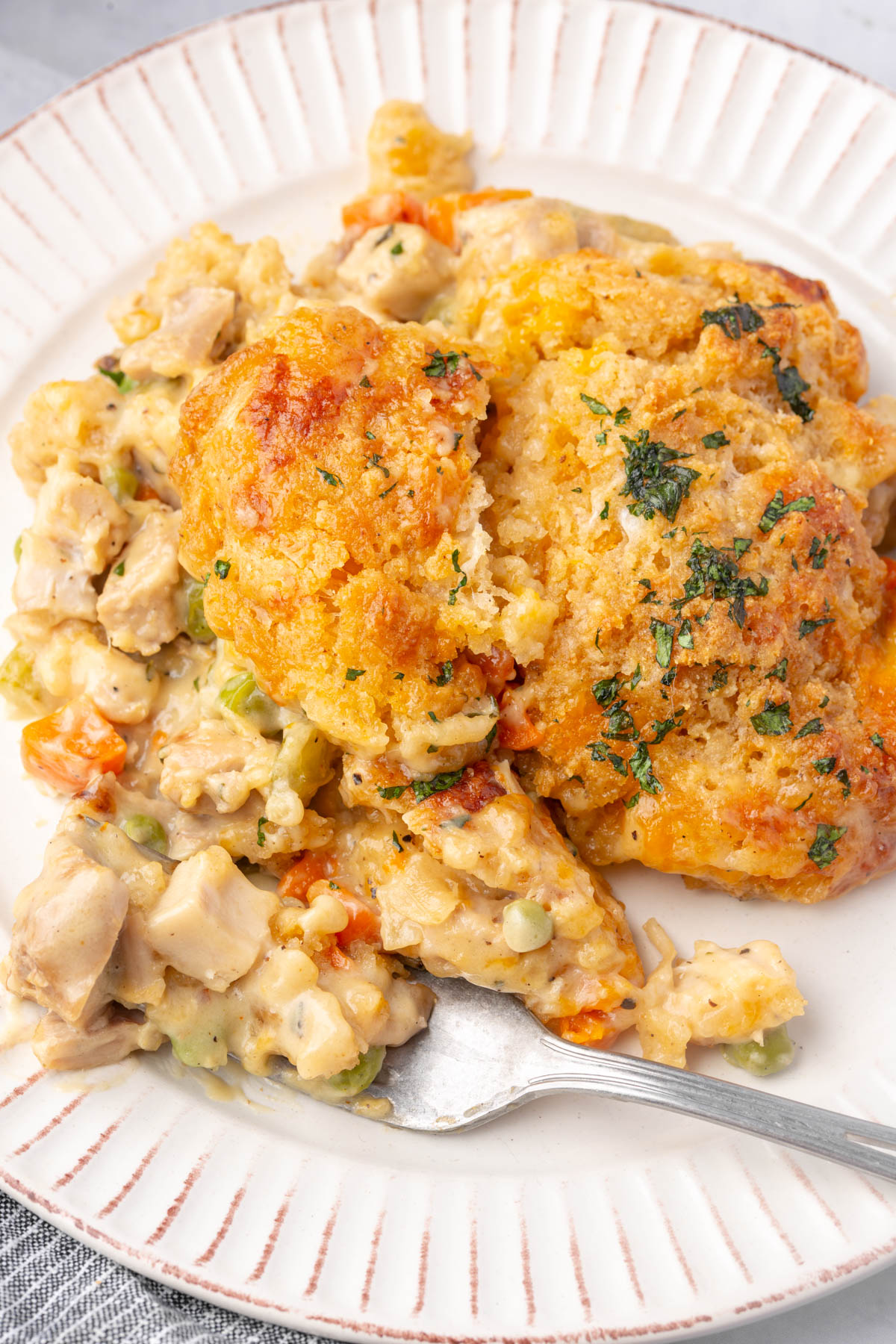 A plate with a serving of creamy chicken and vegetable casserole topped with golden, herb-baked biscuits. A fork rests on the plate, partially embedded in the casserole.