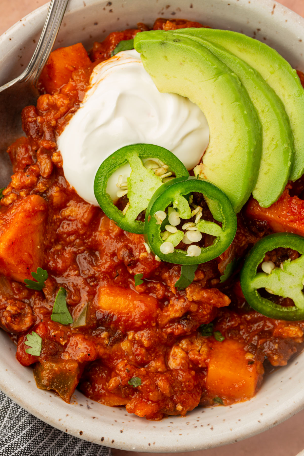 A bowl of chili topped with sliced avocado, jalapeño, and a dollop of sour cream. The chili contains chunks of vegetables and is garnished with chopped herbs. A fork rests on the edge of the bowl.