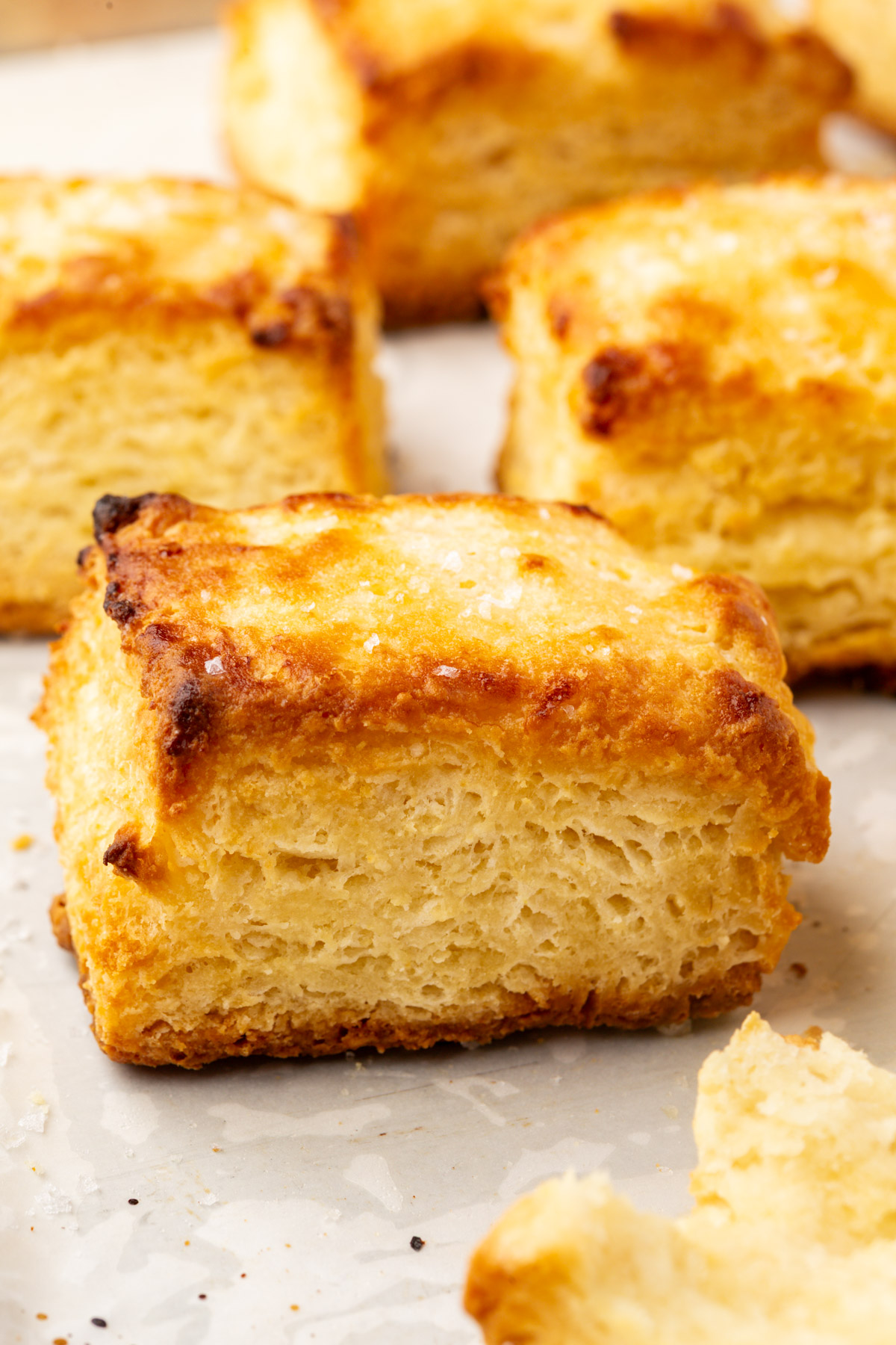 A close-up of golden, flaky buttermilk biscuits on a parchment-lined baking sheet, showing their buttery, crisp edges and soft, layered texture.