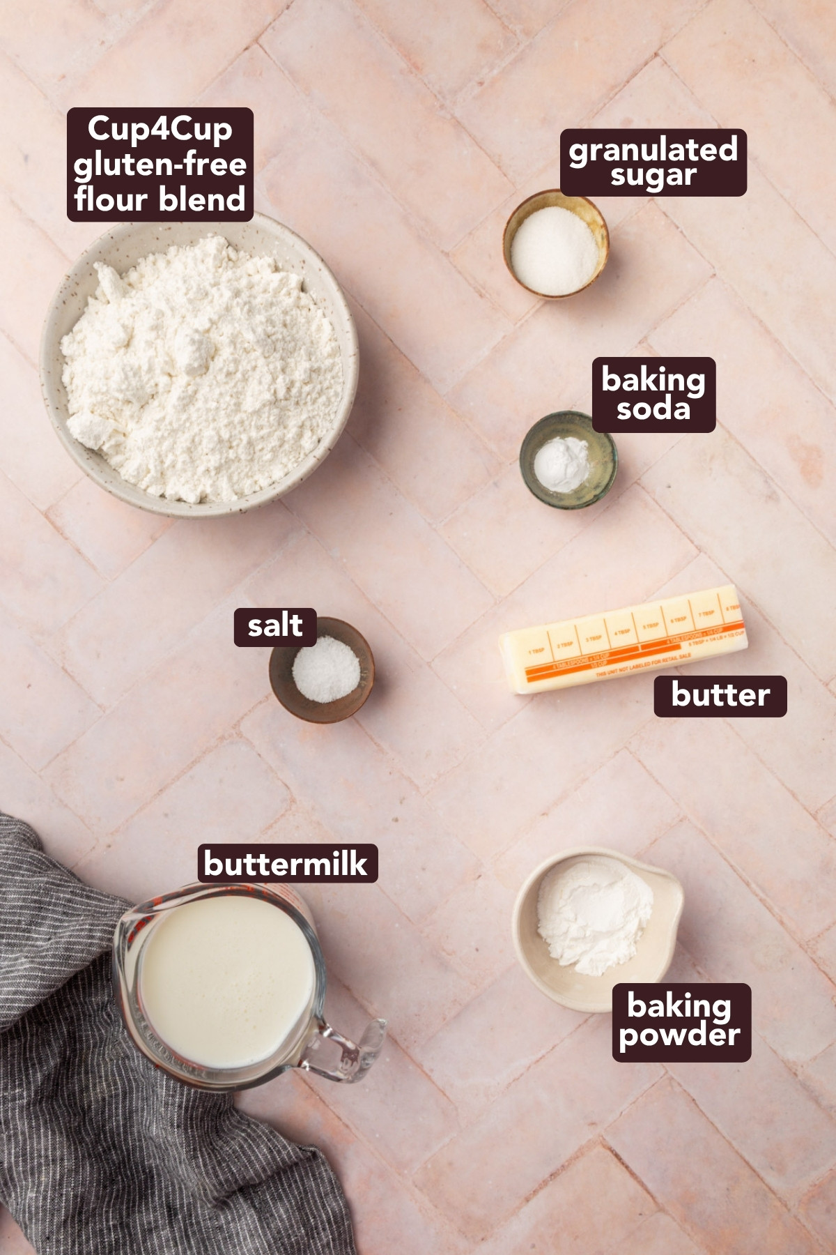 Top-down view of labeled baking ingredients on a pink surface: gluten-free flour blend, granulated sugar, baking soda, salt, butter, buttermilk in a glass jug, and baking powder in small bowls.