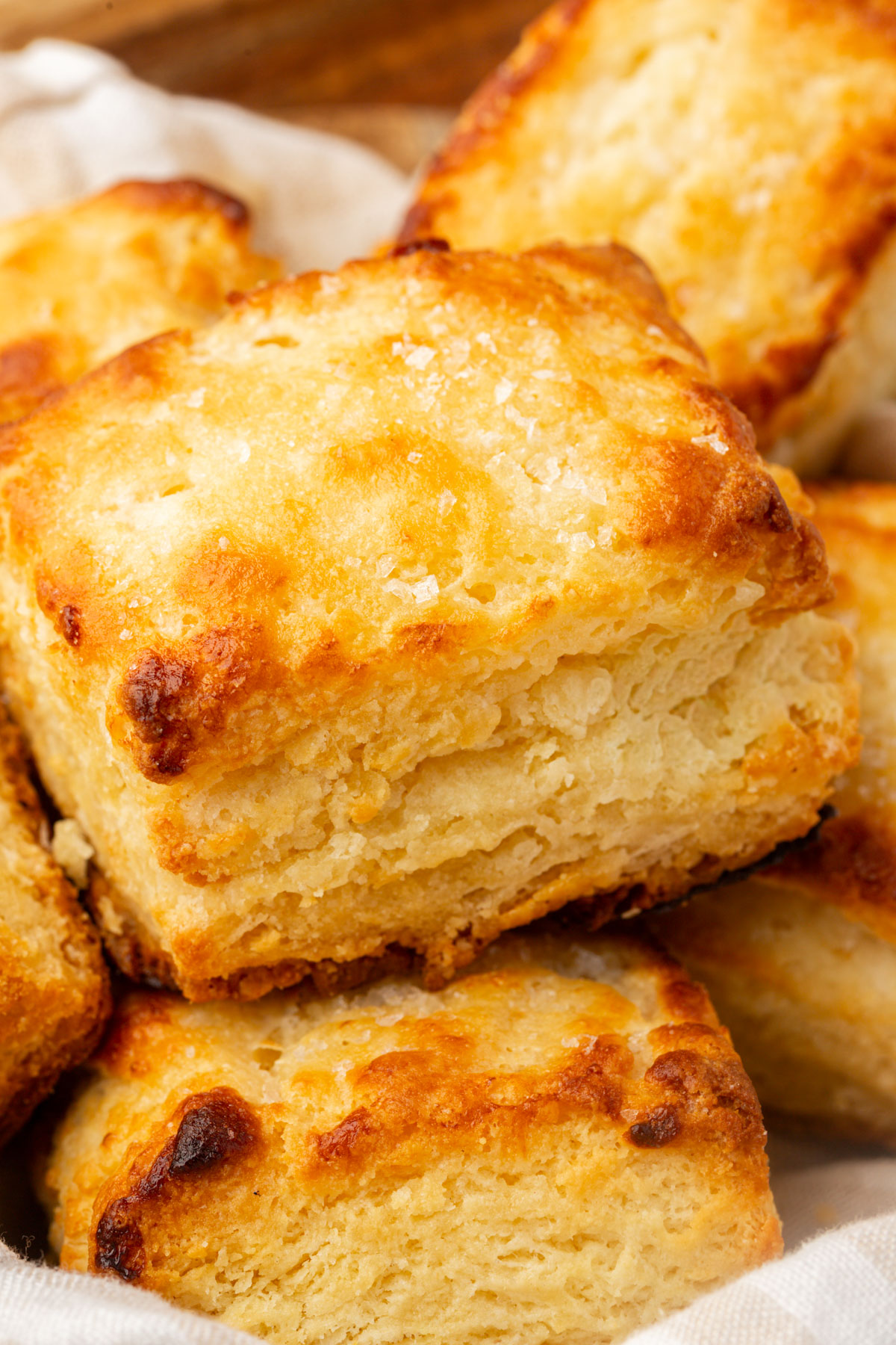 A close-up of golden, flaky biscuits stacked on a white cloth, showing crisp, browned tops and soft, layered interiors.