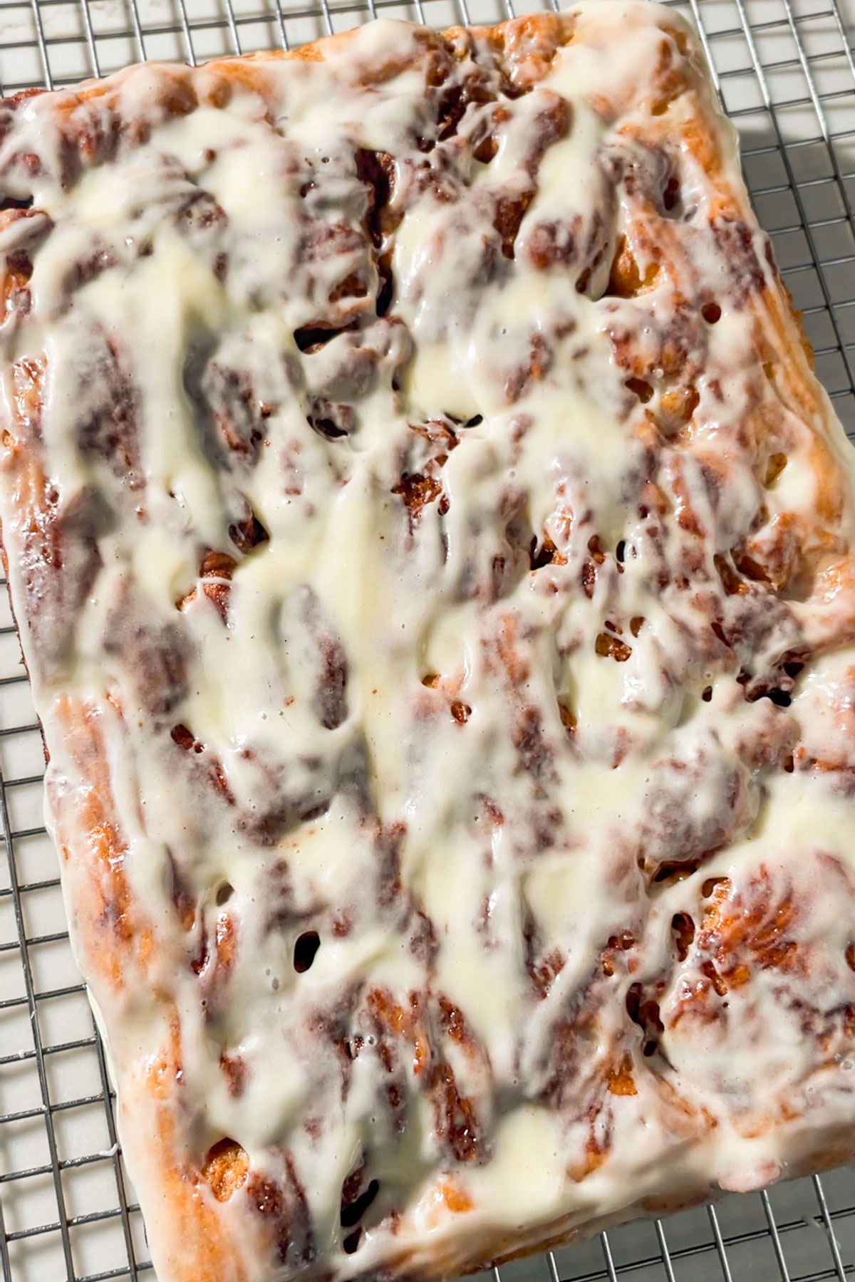 A close-up of a rectangular cinnamon roll focaccia slab topped with creamy white icing, resting on a metal cooling rack. The icing is melting slightly and spread unevenly over the swirled bread.