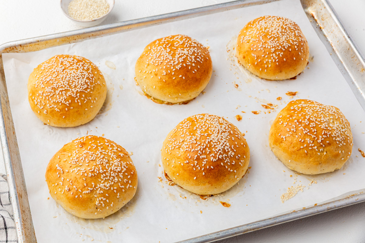 A baking tray lined with parchment paper holds six golden brown, round gluten-free hamburger buns topped with sesame seeds. A small dish of sesame seeds is visible in the background.