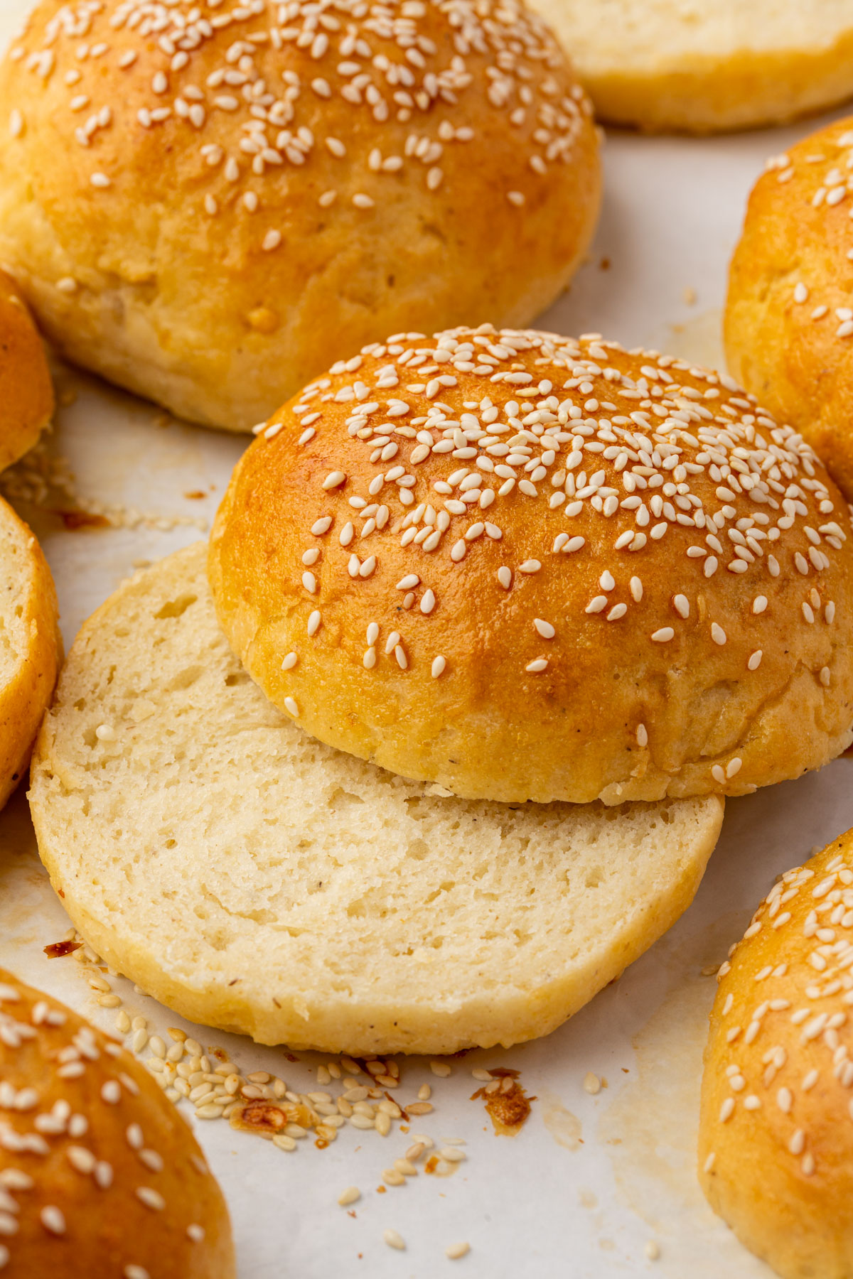 Close-up of several golden brown gluten-free hamburger buns topped with sesame seeds, with one bun sliced open to show its soft interior. The buns are arranged on a light surface.