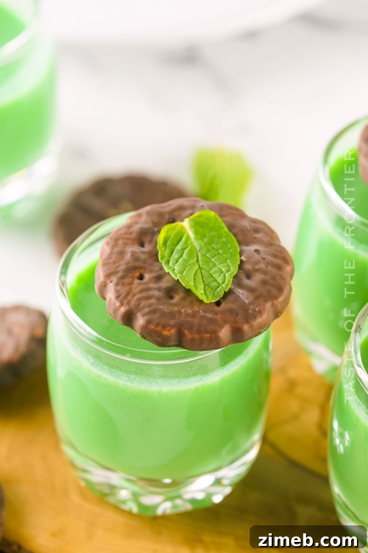 Midnight Thin Mint Shooters 3 A hand holding a shot glass of Dirty Girl Scout Shot, with a Thin Mint cookie on the rim, set against a dark background.