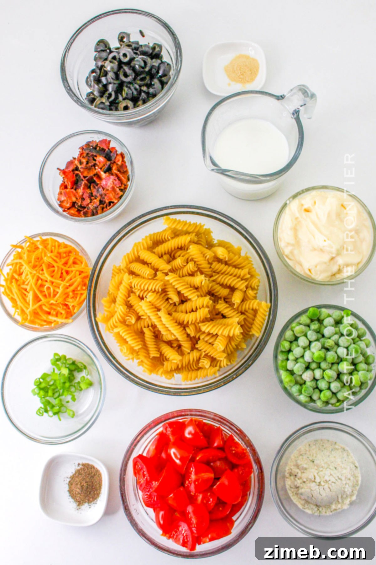 Fresh ingredients for Bacon Ranch Pasta Salad laid out on a cutting board, including pasta, bacon, tomatoes, peas, and cheese.