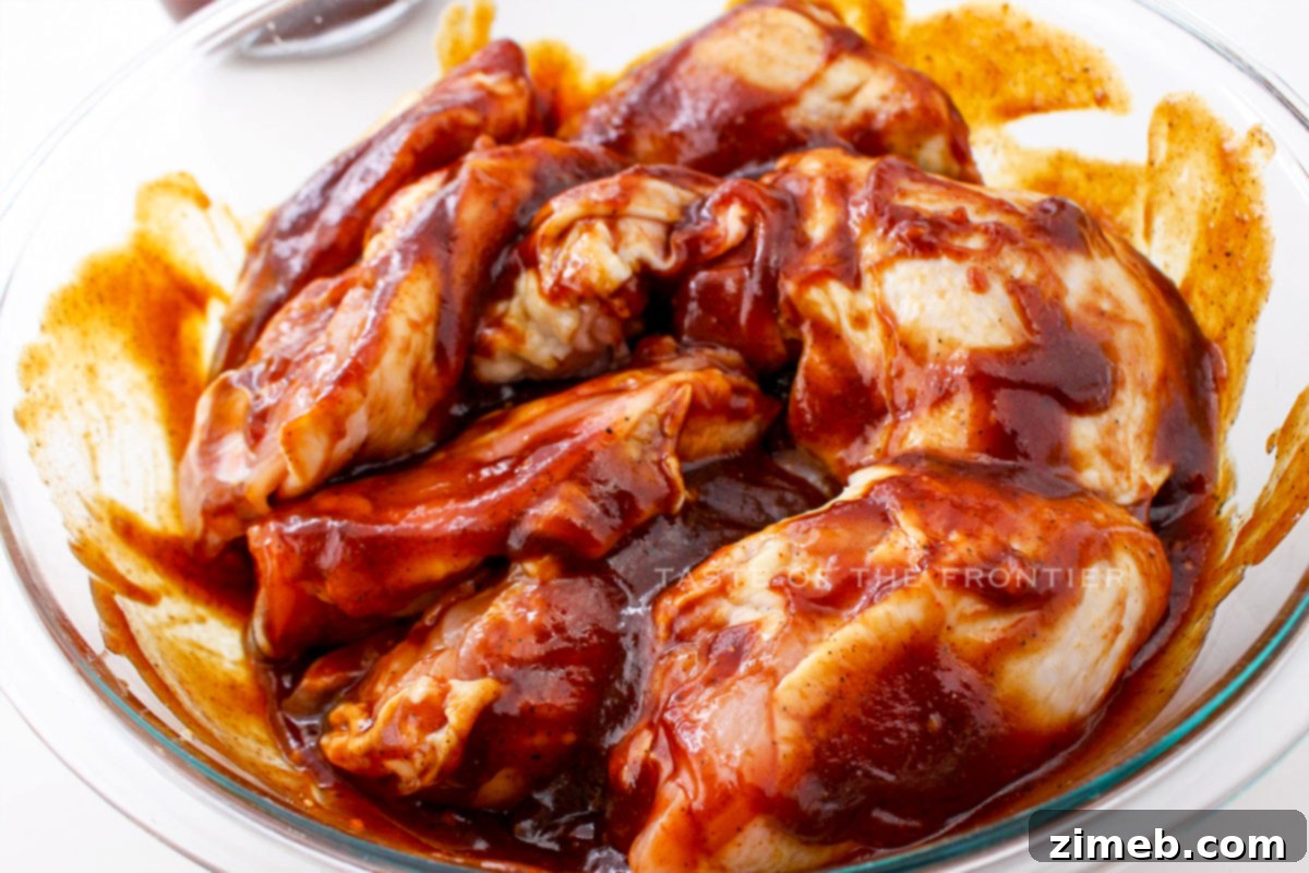 Oven Baked BBQ Chicken Thighs 5 Hands coating raw chicken thighs in a rich BBQ sauce mixture in a bowl, preparing them for baking.