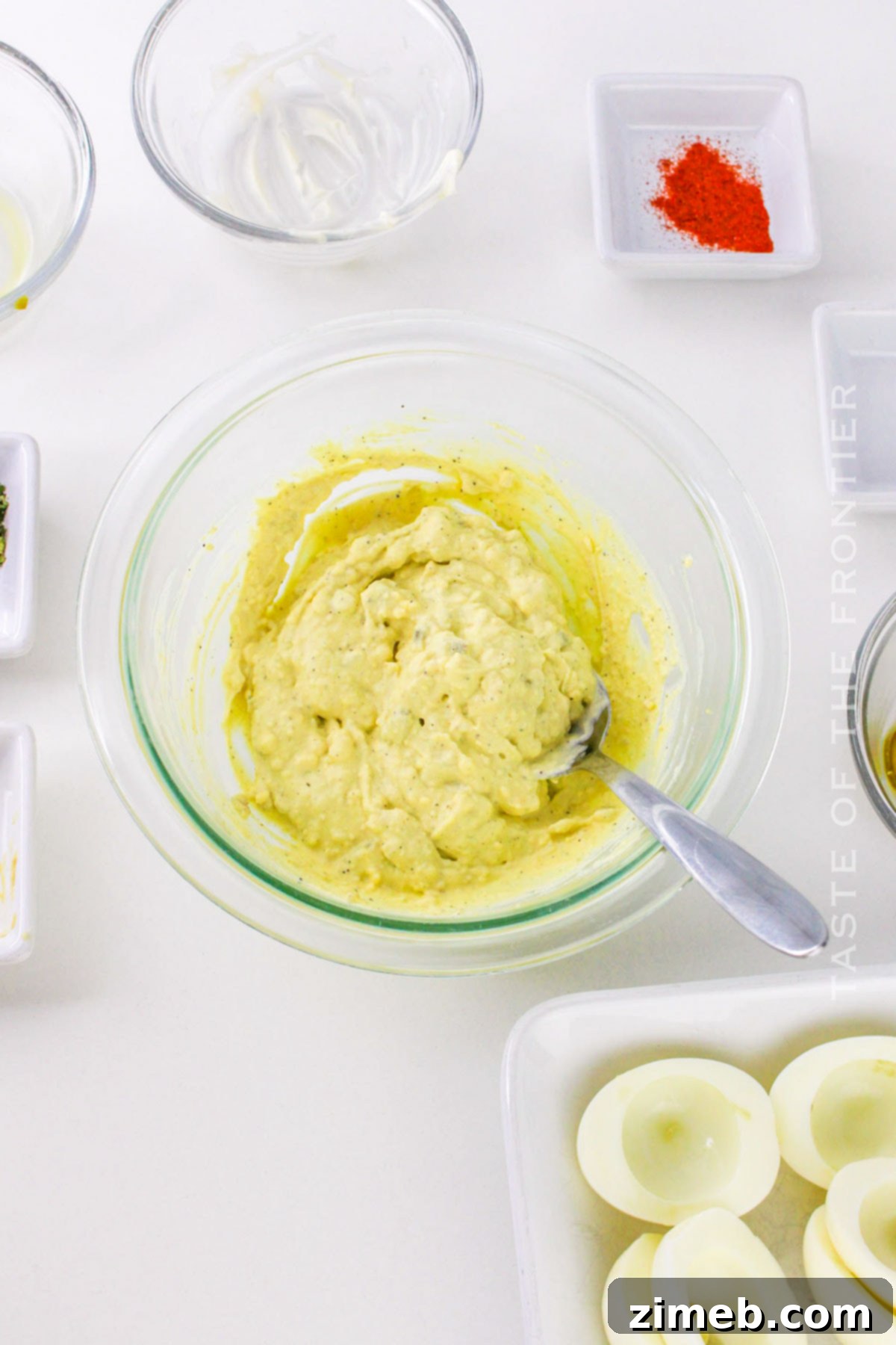 Zesty Jalapeño Deviled Eggs 7 A person mixing the creamy yellow filling for deviled eggs in a bowl with a spatula, surrounded by prepared ingredients.