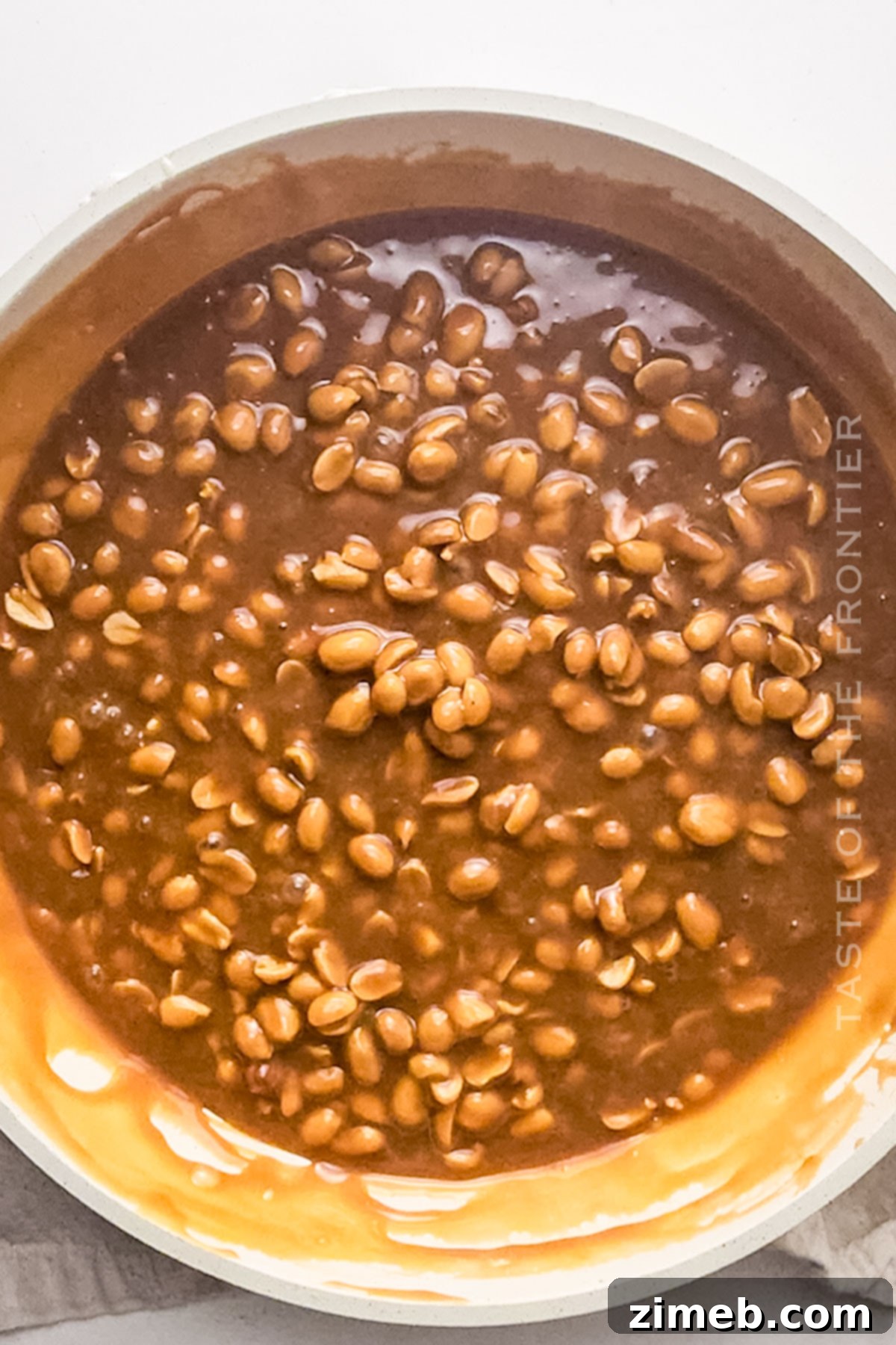 A rich peanut caramel layer being poured over a baked Snickers cheesecake.
