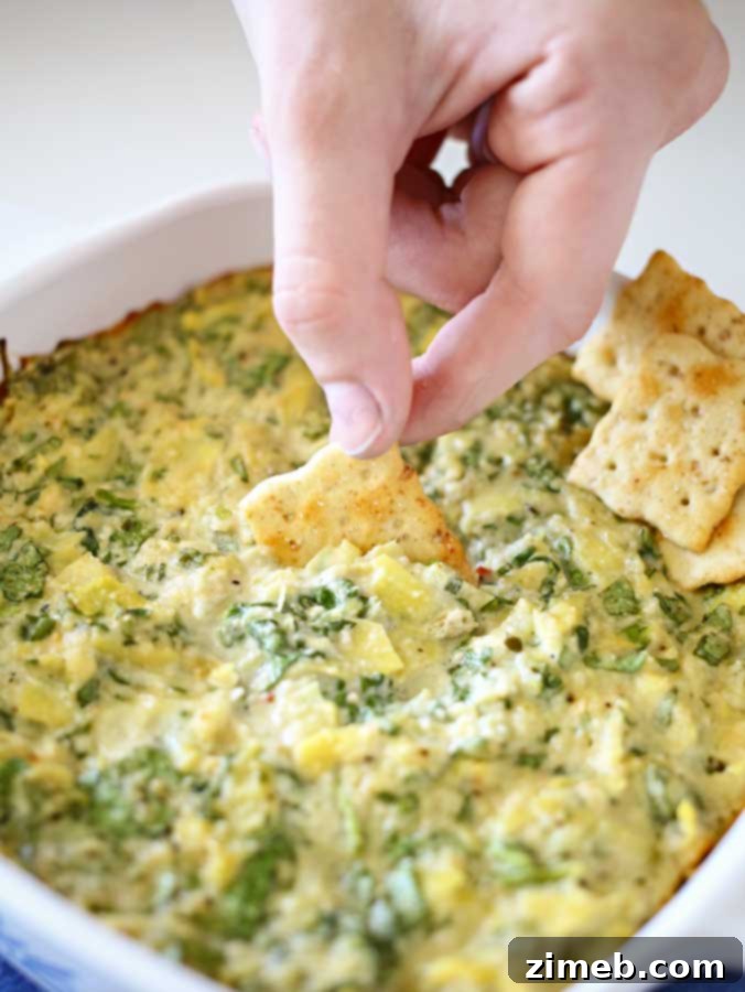 Irresistible Vegan Spinach Artichoke Dip 7 Delicious Artichoke Dip (Vegan) in a small baking dish, served with crackers on a wooden board.