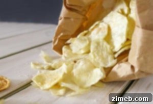 Close-up of homemade potato chips, thinly sliced and golden-brown, spilling from a brown paper bag onto a wooden surface.