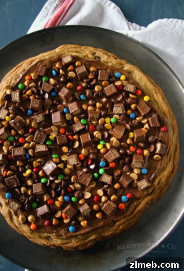 Ultimate Chocolate Chip Cookie Pizza 2 Close-up of a slice of Loaded Chocolate Chip Cookie Pizza, showing the gooey melted chocolate, peanut butter, and butterscotch topping, with crunchy Kit Kat pieces and colorful M&M's on a thick cookie base.